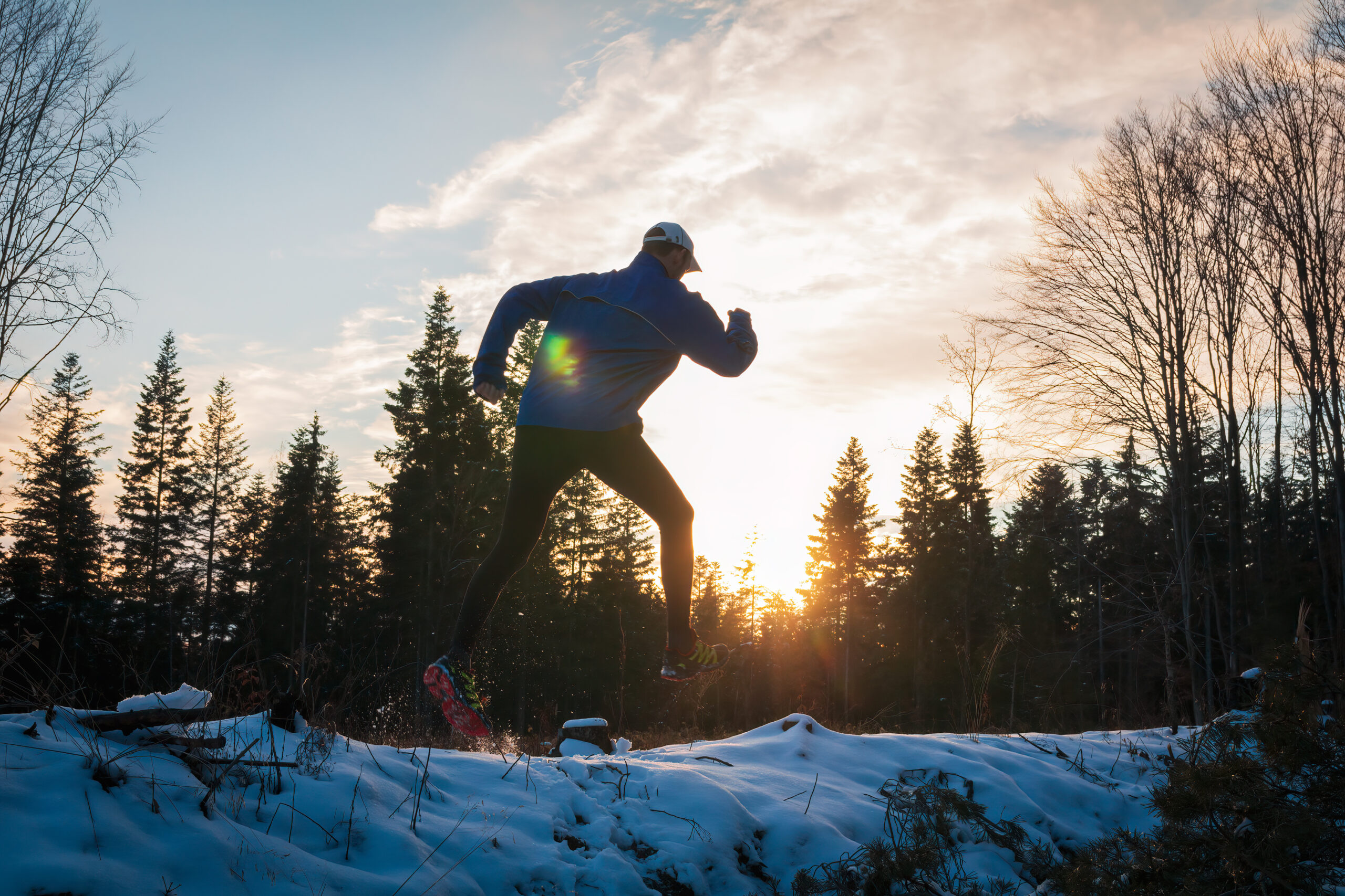 Young caucasian man running on snow in Czech winter landscape at sunset. Sport athlete in jacket, leggings and cap