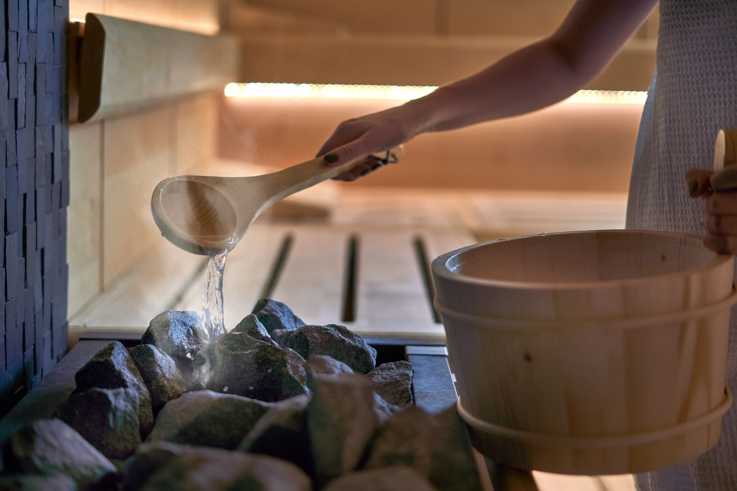 Woman in sauna pouring water to hot stones to produce steam Woman in sauna - real, authentic moment. Women pouring water to hot stones to produce steam.